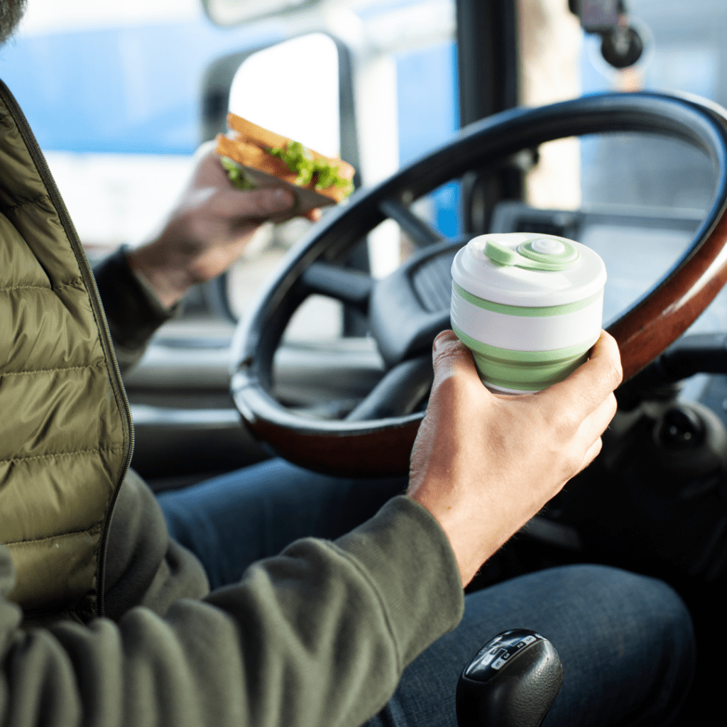 En la imagen se muestra un hombre sosteniendo un sándwich y un termo de café, sentado frente al volante de un camión