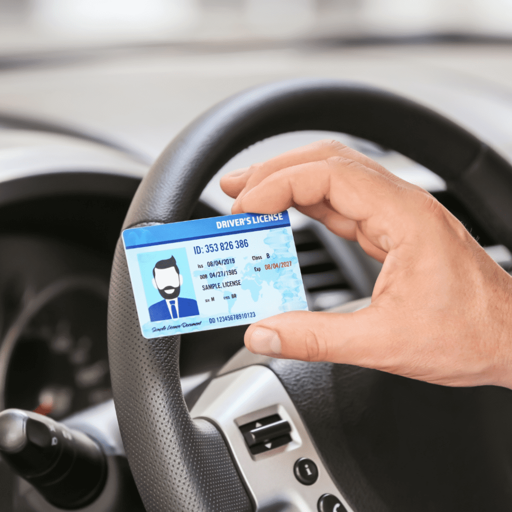En la imagen se muestra una mano sosteniendo una licencia de conducir, frente a un volante de auto