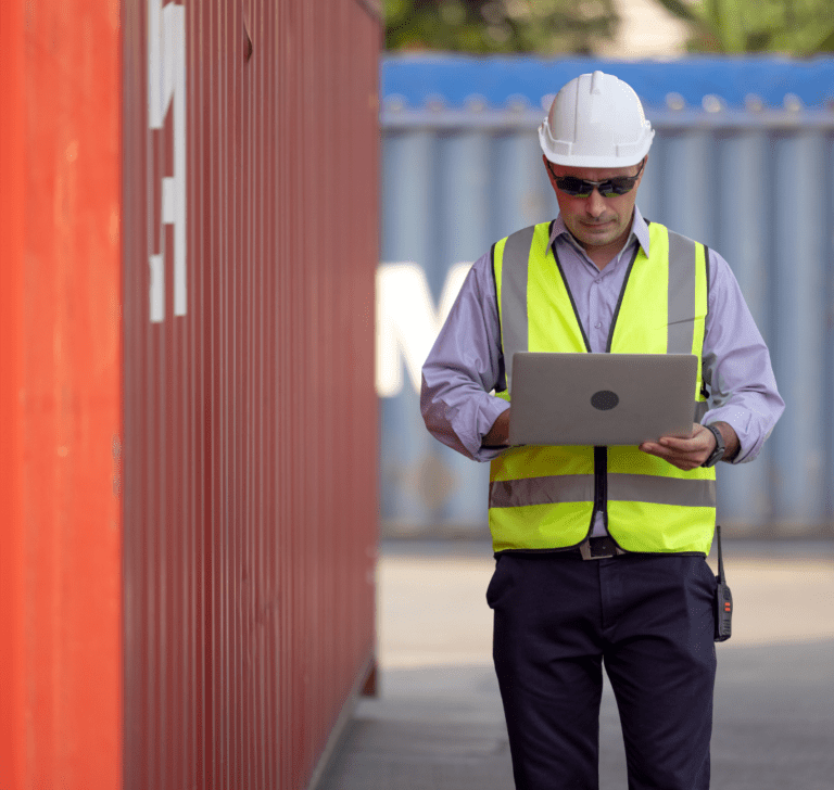 En la imagen se muestra un hombre con una computadora, junto a contenedores de carga