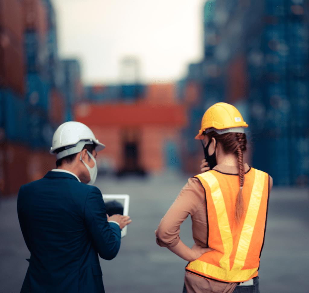 En la imagen se muestra un hombre con una computadora, junto a una mujer, en un área con contenedores de carga